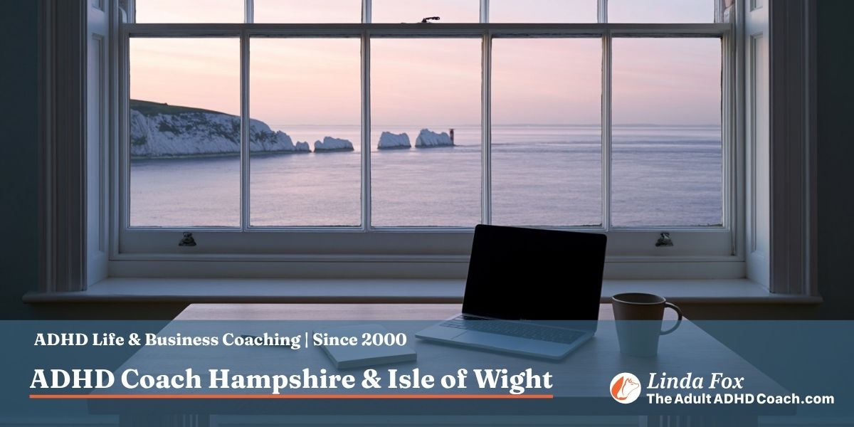 A neat home study desk in an Isle of Wight clifftop cottage at first light, with a sash window framing The Needles chalk stacks and lighthouse in the soft dawn beyond
