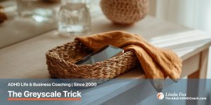 Blog post: The Greyscale Trick — a smartphone resting in a small woven basket on a console table, representing parking the phone and reducing mindless scrolling