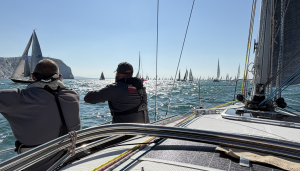 Two crew members on a yacht during the Round the Island Race, looking out at the fleet near the Needles. Bright sunlight scatters off the water and dozens of sails stretch to the horizon.