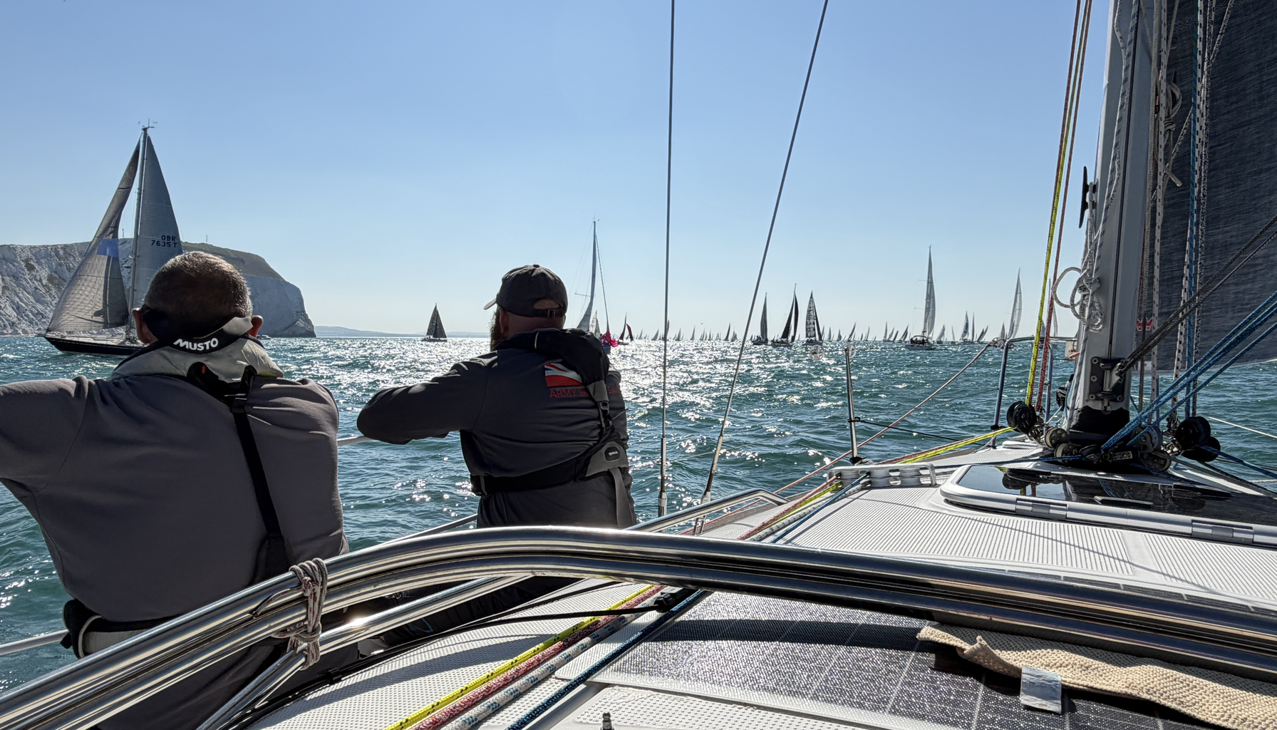 Two crew members on a yacht during the Round the Island Race, looking out at the fleet near the Needles. Bright sunlight scatters off the water and dozens of sails stretch to the horizon.