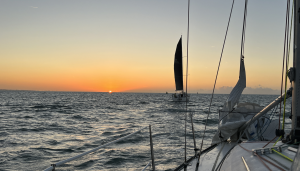 The view from a yacht at sunset. The sun sits low on the horizon over open water, one boat sails ahead under a dark sail, and the guardrail and foredeck run along the bottom of the frame.