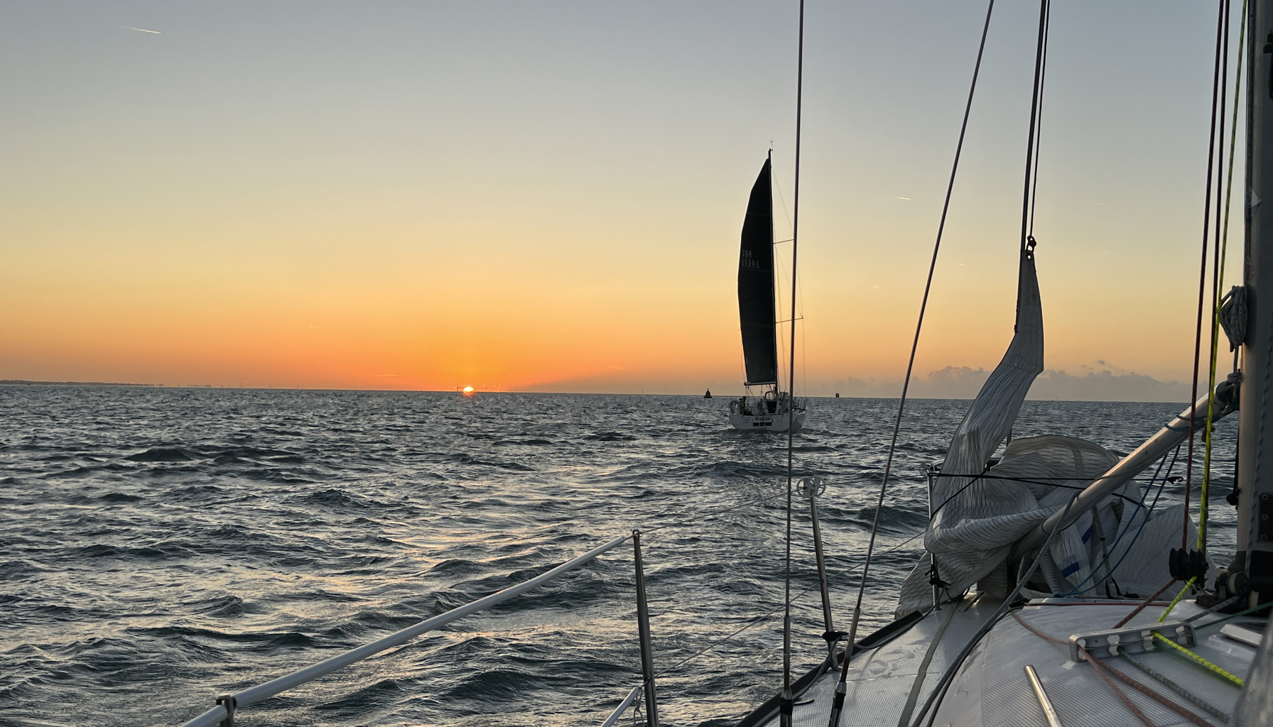 The view from a yacht at sunset. The sun sits low on the horizon over open water, one boat sails ahead under a dark sail, and the guardrail and foredeck run along the bottom of the frame.