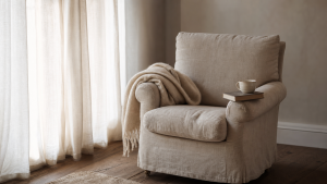 A worn upholstered cream linen armchair beside a window, cream wool throw draped over one arm, a closed hardback book and small half-empty ceramic cup on the other arm, soft morning light through gauzy curtains.