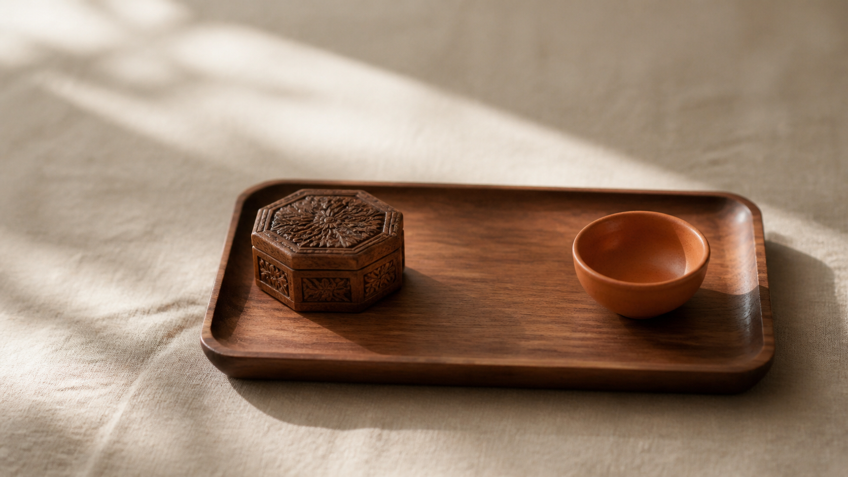 A small wooden tray on warm cream linen holds a carved wooden box and a small terracotta ceramic bowl in soft morning light.